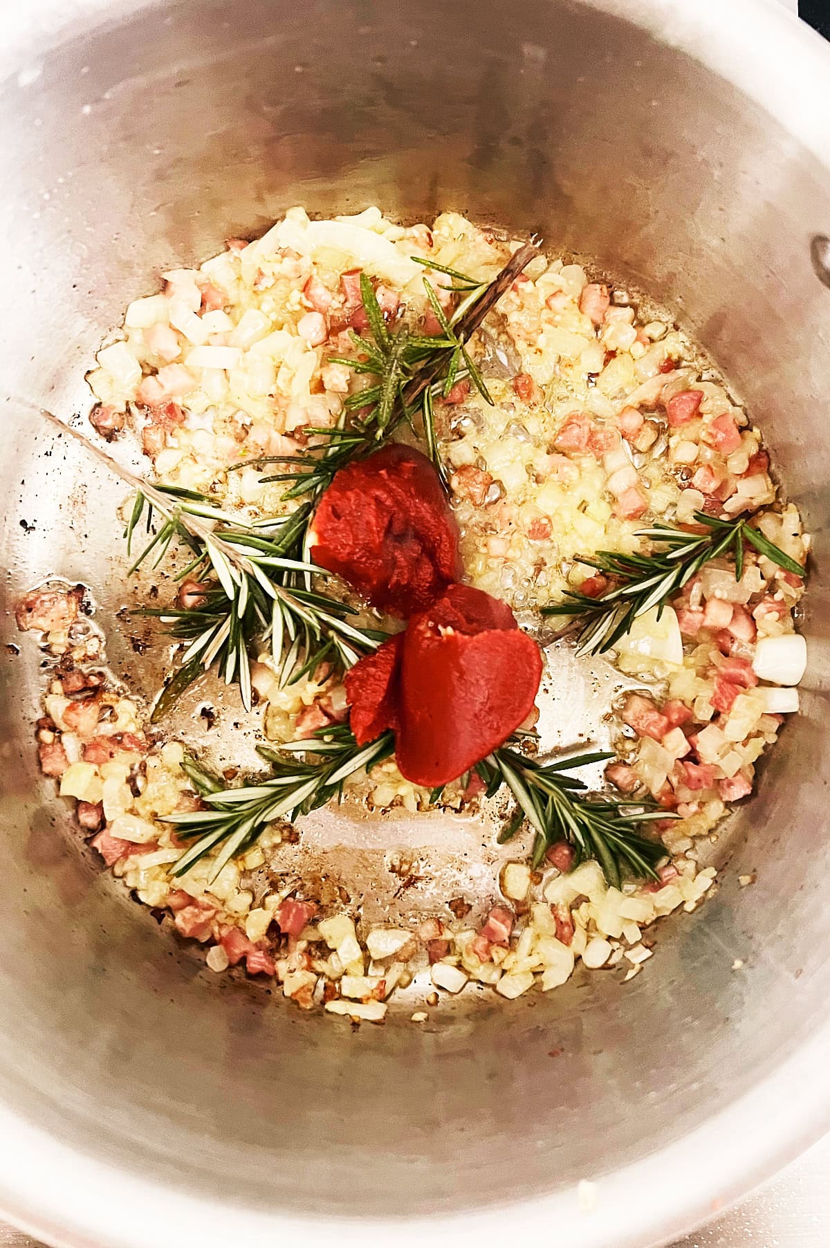 Chopped onions and diced pancetta cooking in a pot with sprigs of rosemary and a dollop of tomato paste on top.