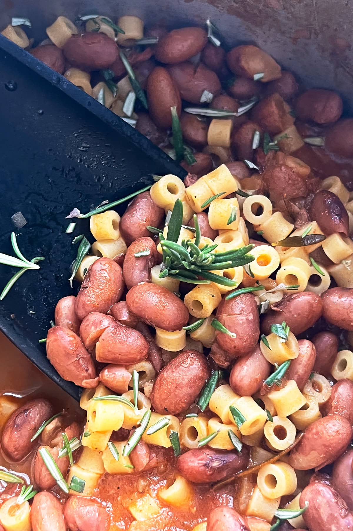 A close-up of a saucy pot filled with cooked red beans, small pasta, and garnished with fresh rosemary and a black ladle.