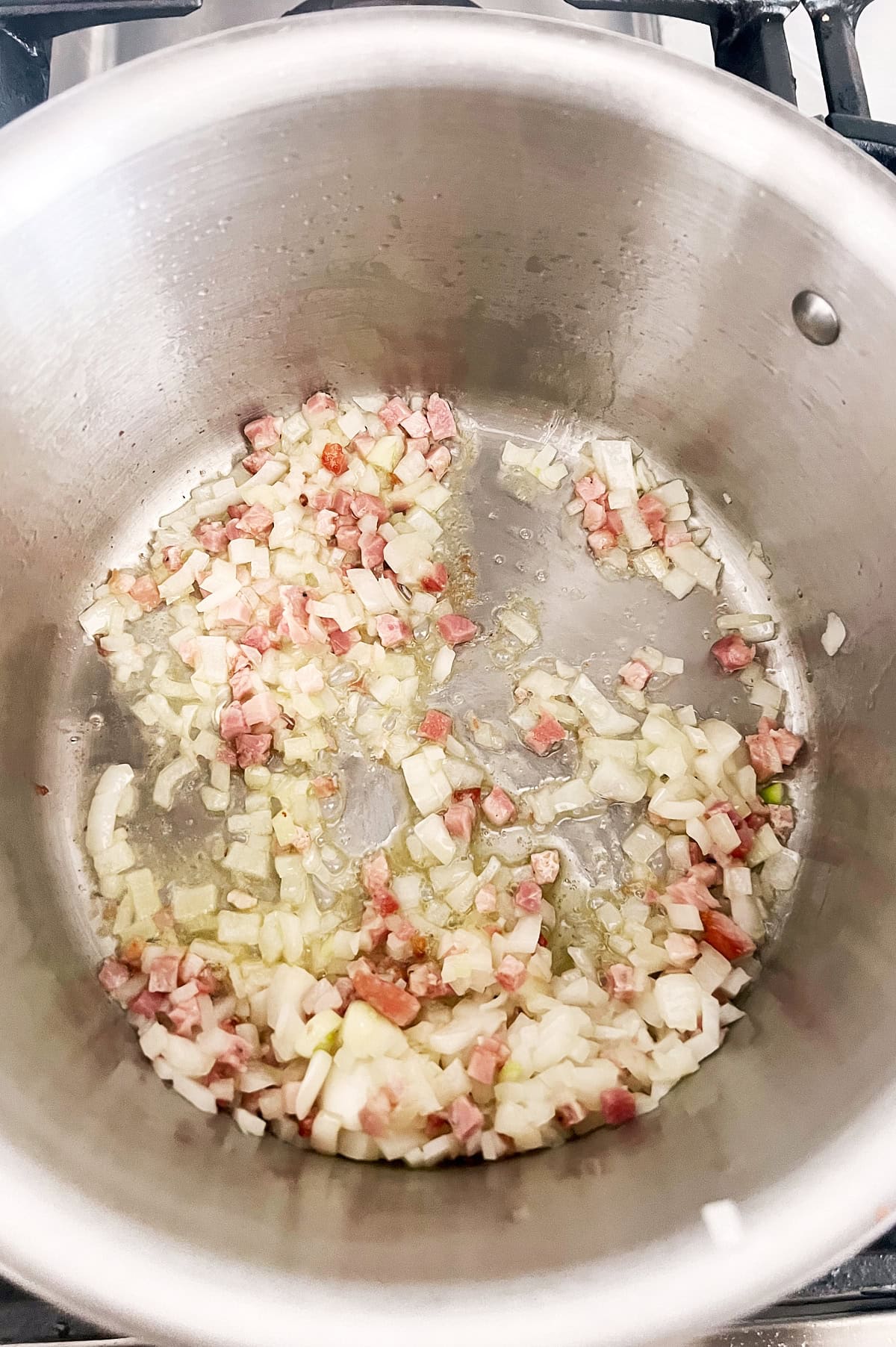 Chopped onions and diced pancetta are being sautéed in a stainless steel pot on a stove, with a light coating of oil visible.
