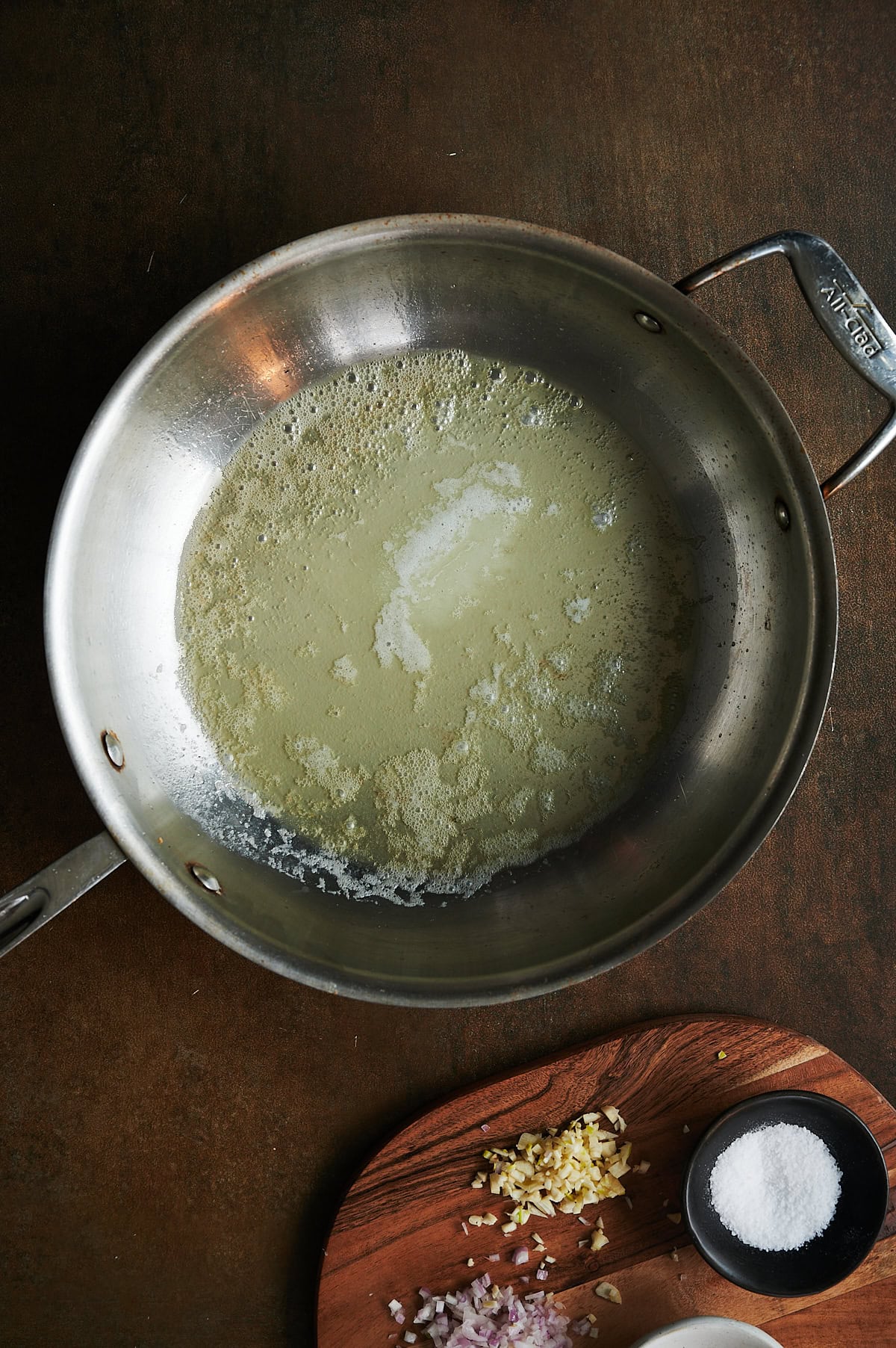 Skillet of melted butter on a brown background with board of shallots and garlic nearby.