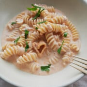 A white bowl of pink pasta sauce rotini and a heart noodle with a fork, parmesan cheese and parsley.