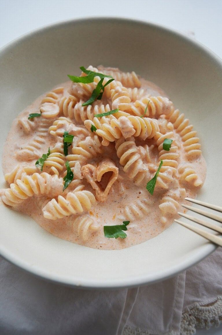 A white bowl of pink pasta sauce rotini and a heart noodle with a fork, parmesan cheese and parsley.