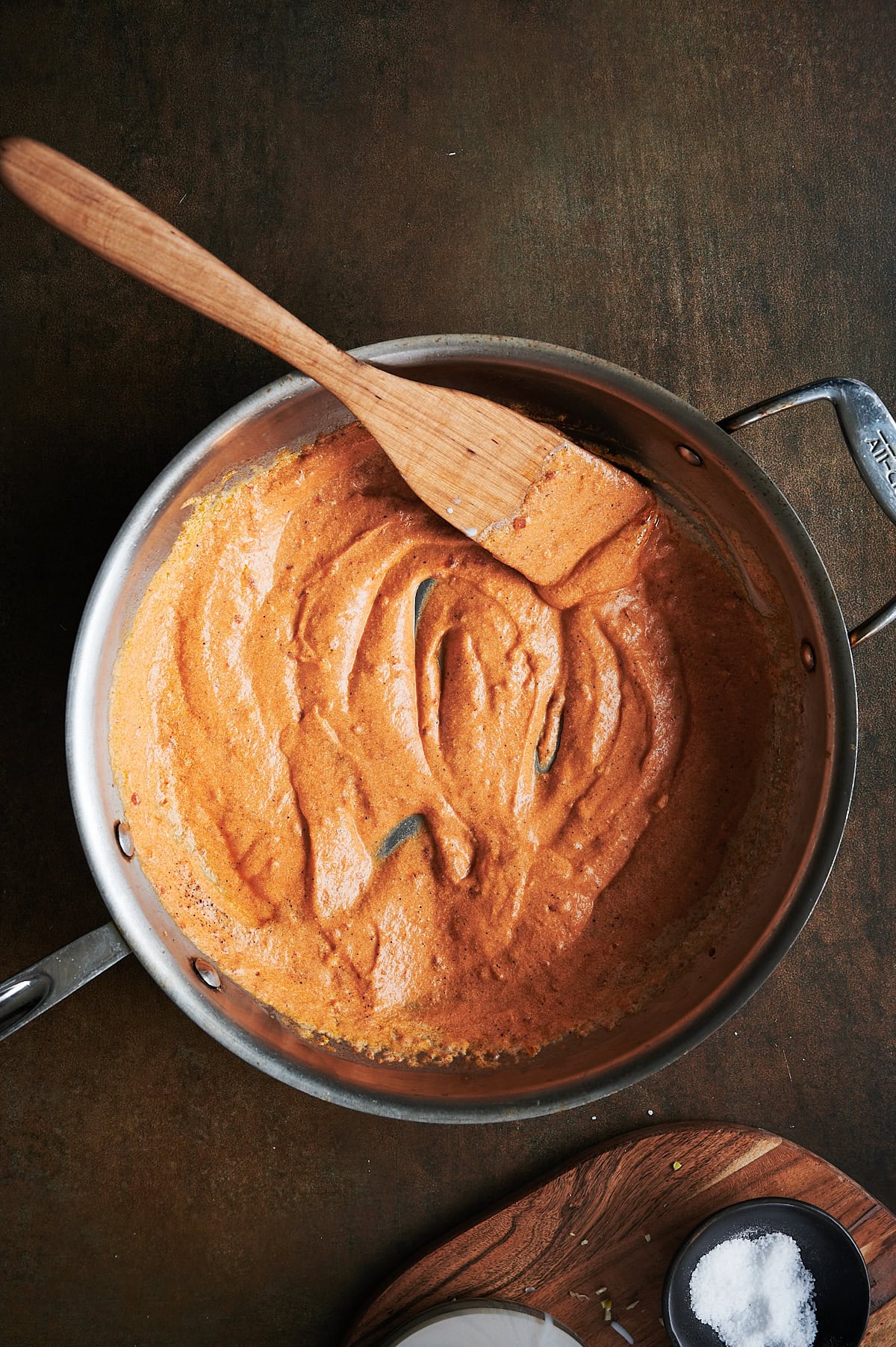 Orange colored tomato cream sauce being stirred by wooden paddle in a stainless skillet on a brown background.