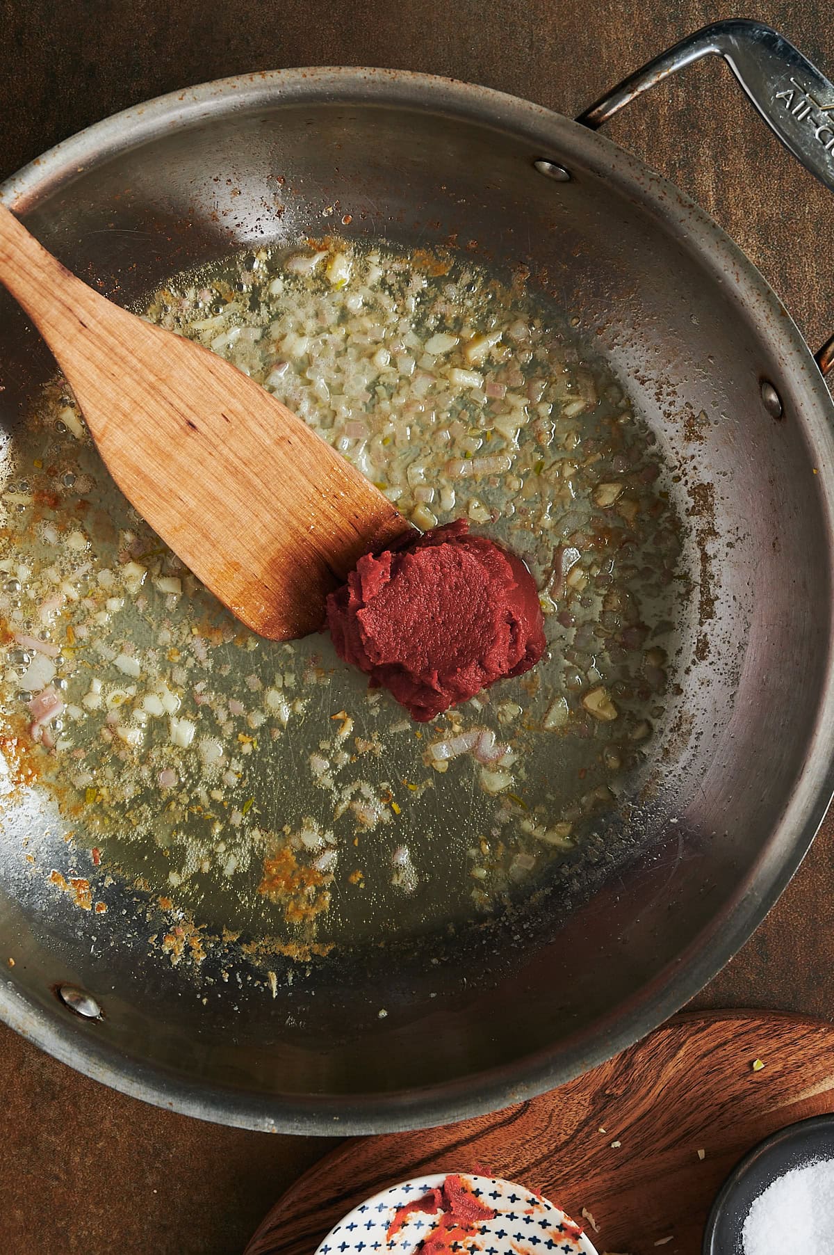 Wooden paddle stirring tomato paste into a skillet of sauteed onions.