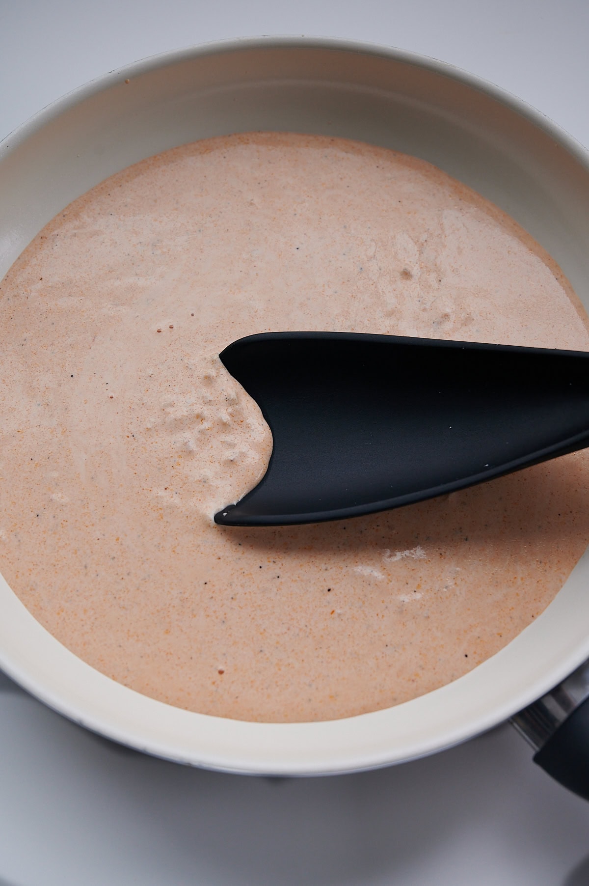 Black silicon spoon stirring cream into pink pasta sauce in a white skillet.