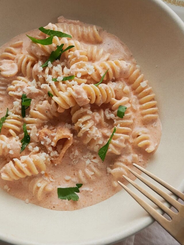 A bowl of fusilli pasta is coated in a creamy tomato sauce and sprinkled with grated cheese. Fresh green herbs adorn the dish, and a fork rests on the side.