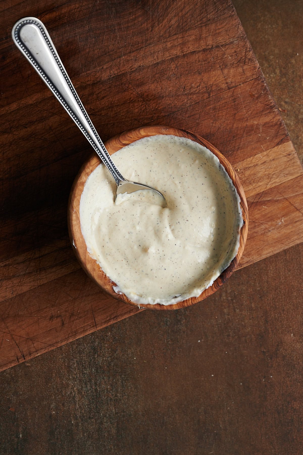 A small wooden bowl of creamy white sauce with visible black specks, placed on a wooden cutting board.