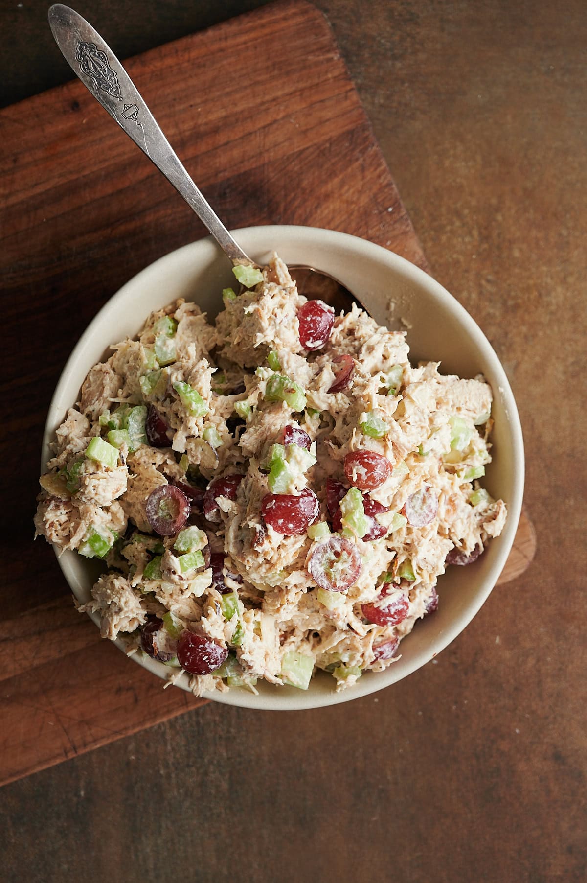 A bowl of chicken salad with sliced red grapes and chopped celery and a spoon sits on a wooden cutting board.