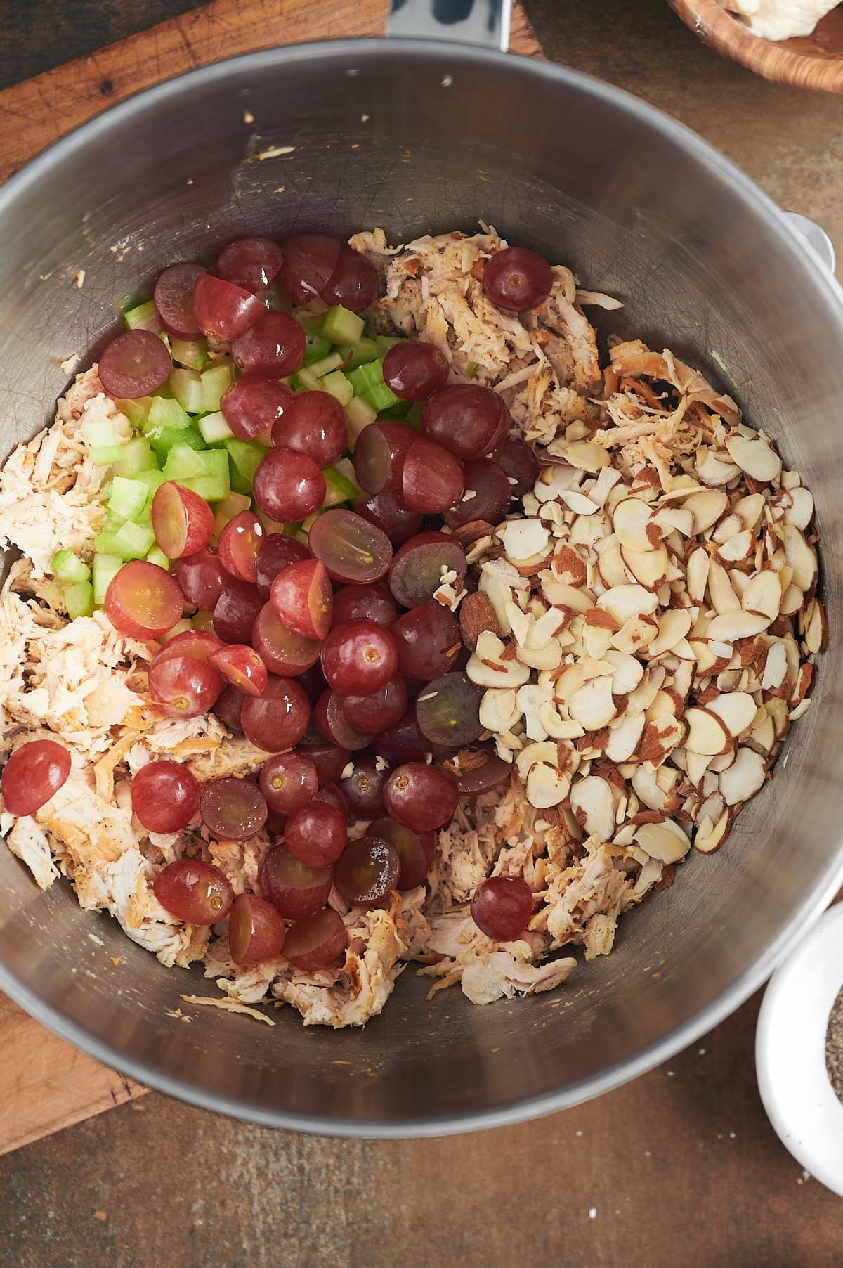 A stainless steel mixing bowl contains shredded chicken, halved red grapes, sliced celery, and sliced almonds, each in separate sections, ready to be mixed together.