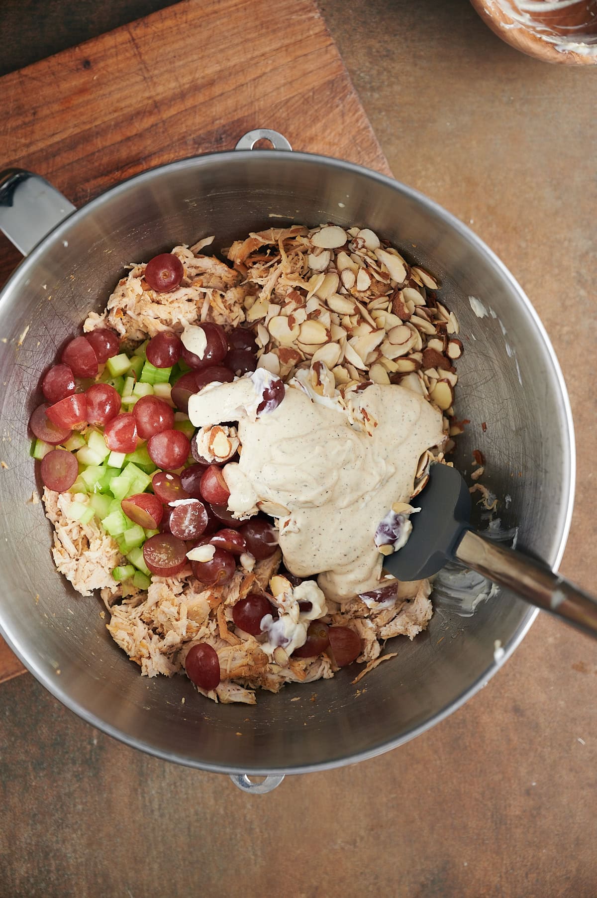 A metal mixing bowl contains shredded chicken, sliced grapes, chopped celery, sliced almonds, and a dollop of creamy dressing being mixed by a black spatula.