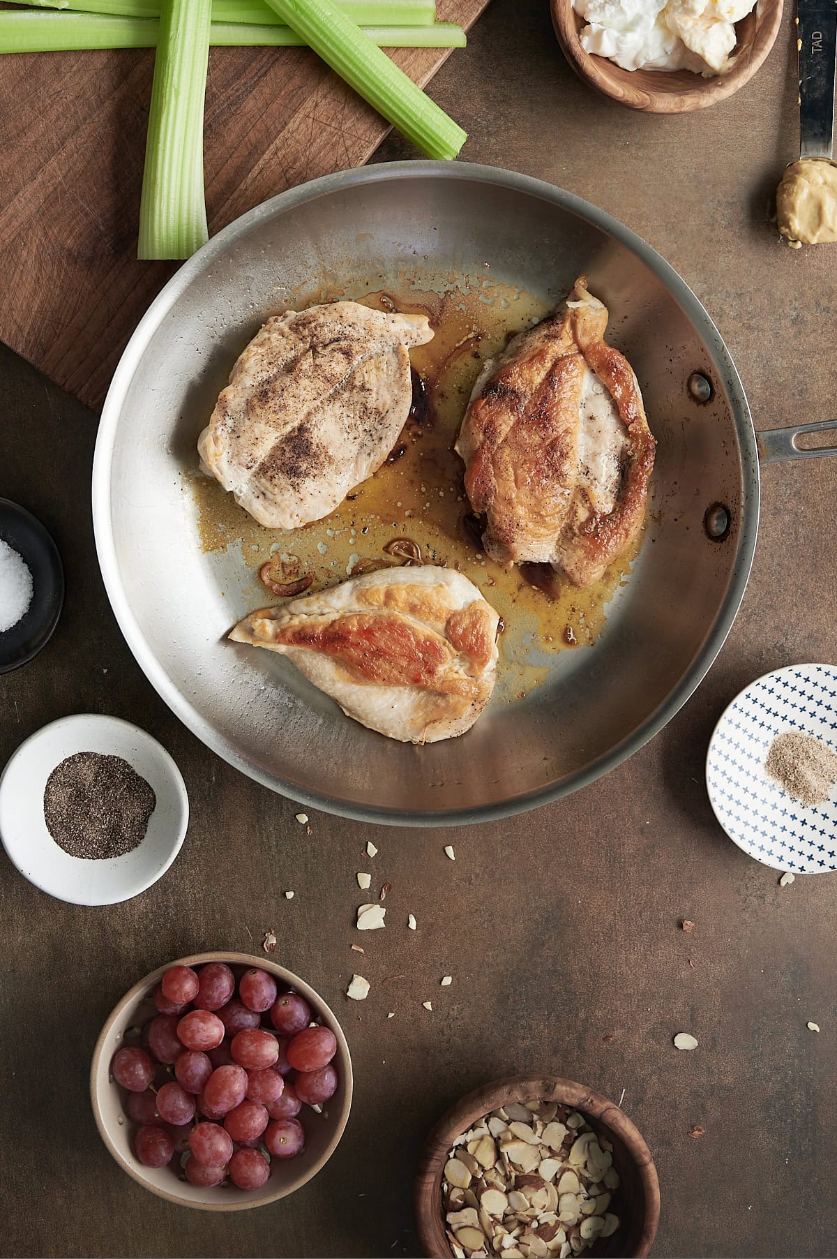 A stainless steel pan with three cooked chicken breasts sits on a brown surface, surrounded by bowls of grapes, sliced almonds, ground pepper, celery stalks, salt, and other ingredients.