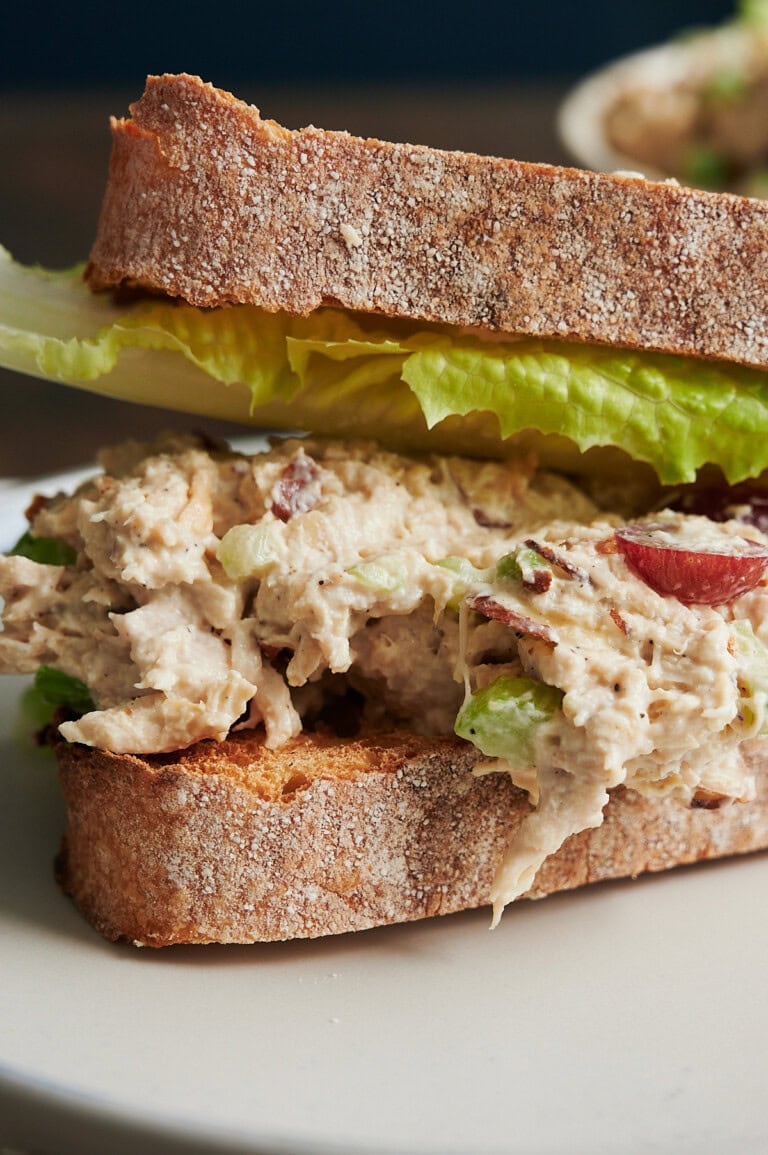 A close-up of a chicken salad sandwich on crusty bread with lettuce. The chicken salad contains pieces of grape and appears creamy.