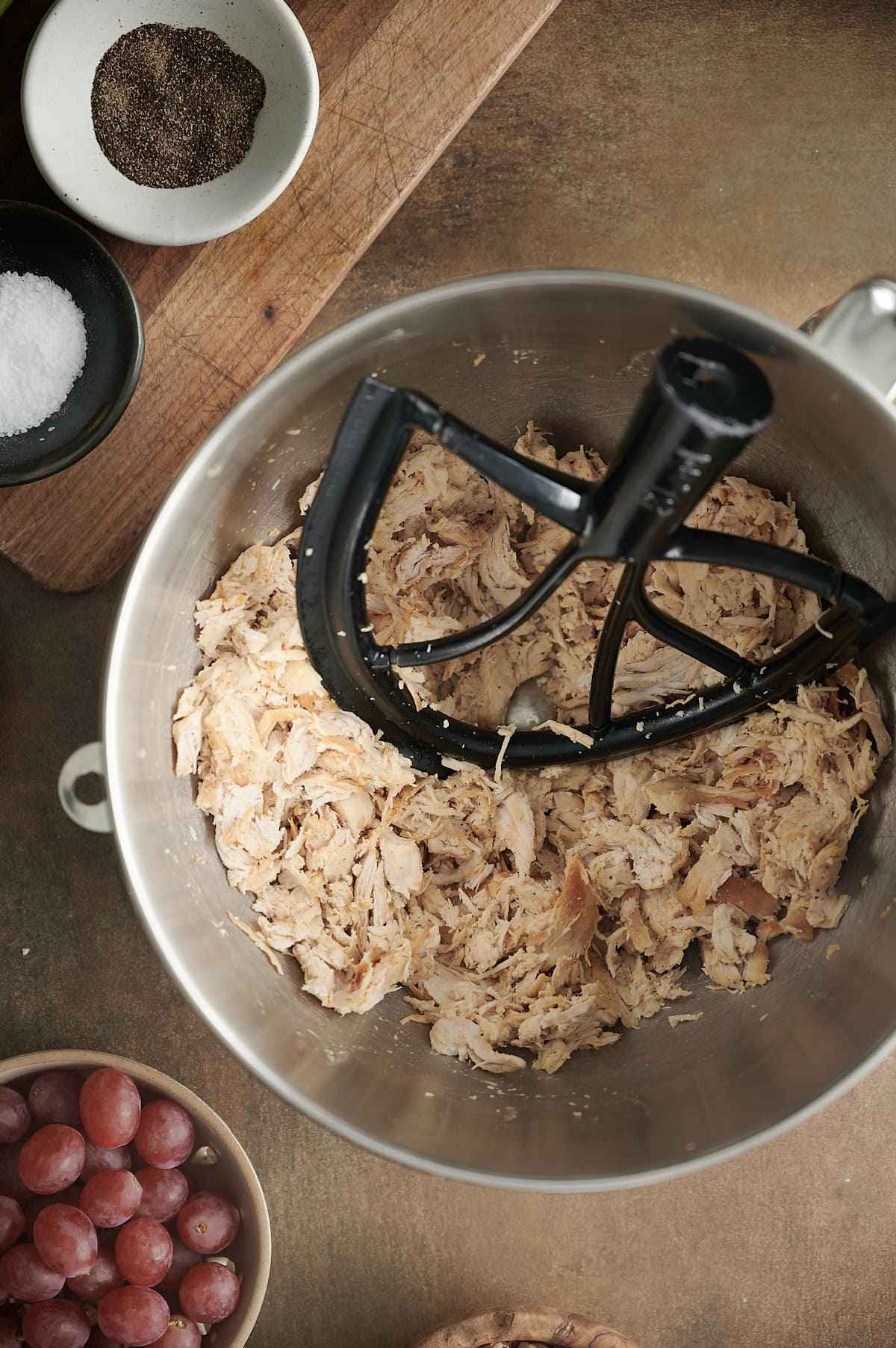 A metal mixing bowl contains shredded chicken with a paddle attachment. Surrounding the bowl are bowls of salt, pepper, and red grapes, all on a wooden surface.