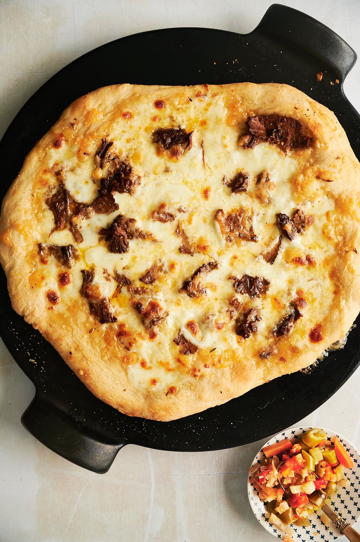 A cooked pizza with melted cheese and pieces of Italian beef on a round black pizza pan, next to a small dish containing giardiniera.