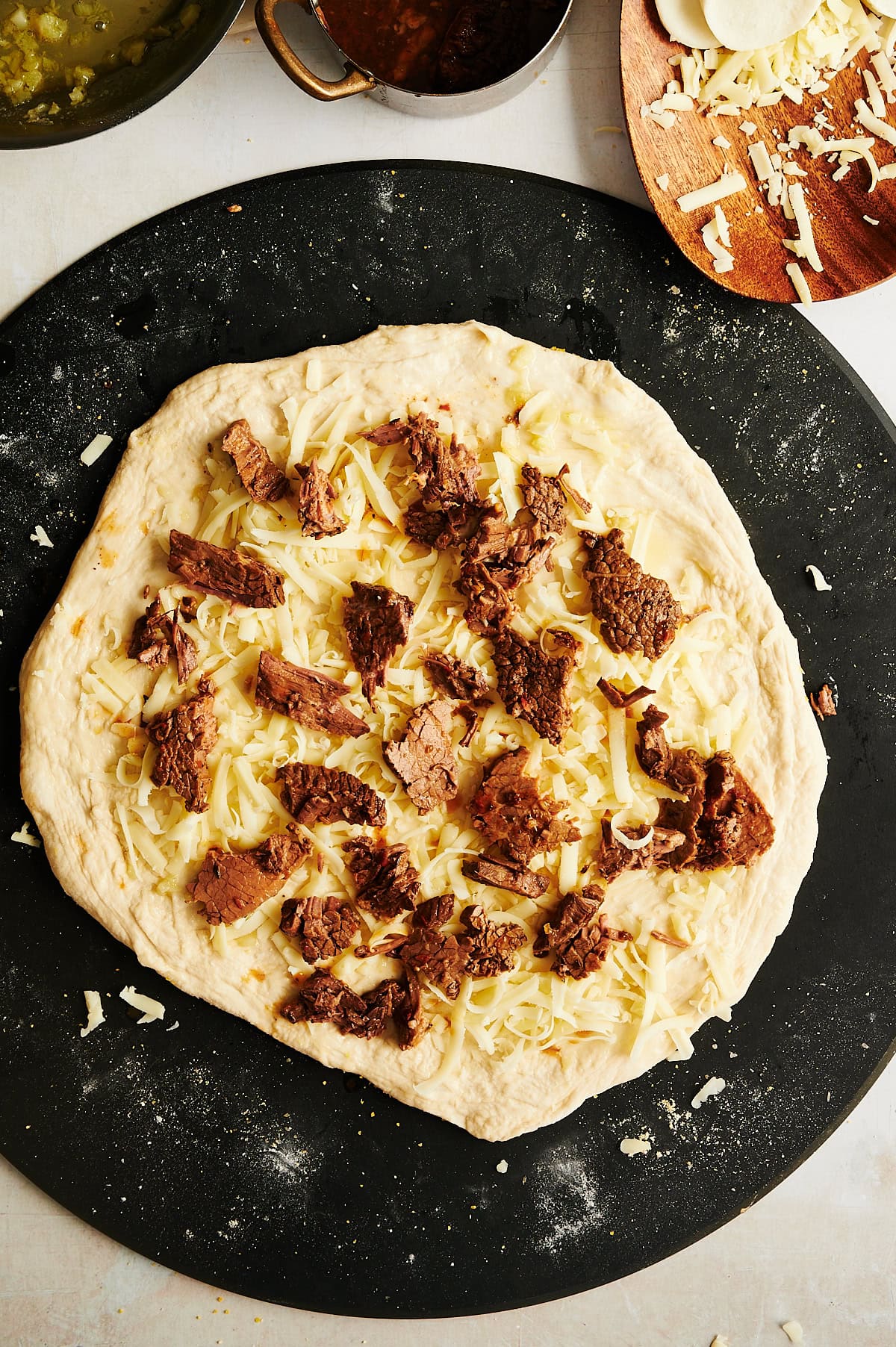 Unbaked pizza dough on a dark surface topped with shredded cheese and chunks of cooked beef.