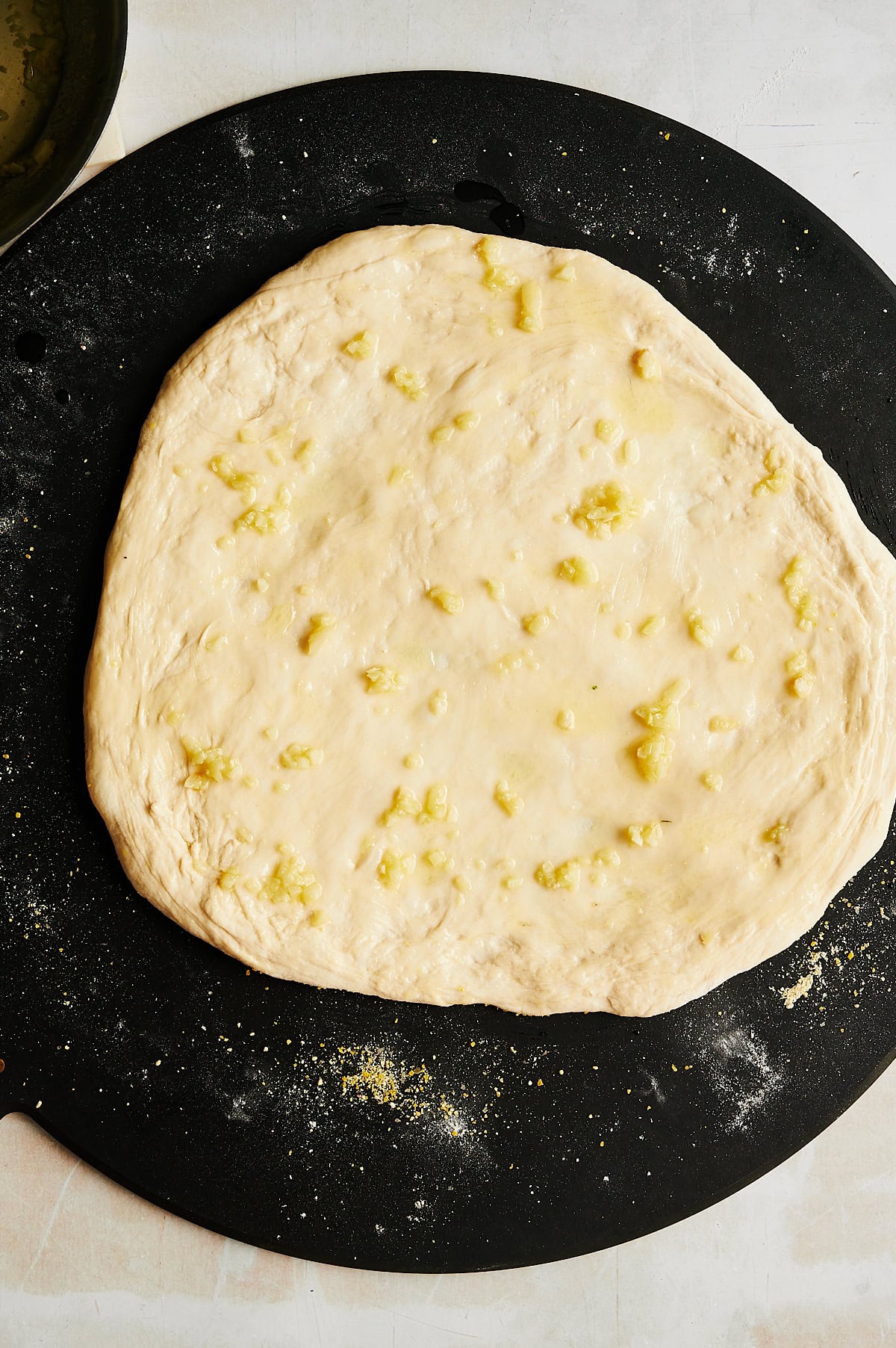 A round, uncooked pizza dough is spread out on a black surface and brushed with olive oil and small pieces of garlic.
