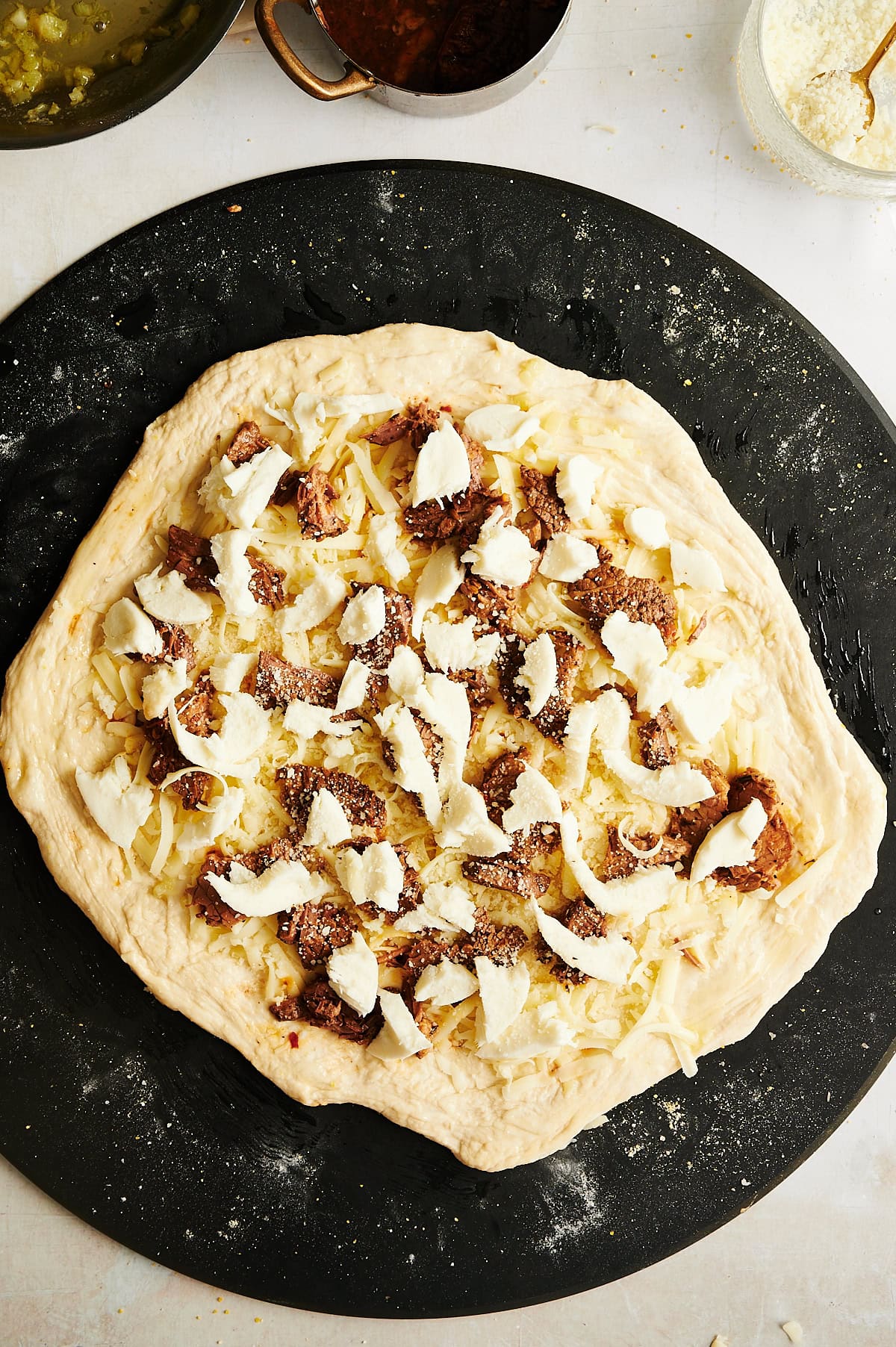 Unbaked pizza on a black surface, topped with chunks of cheese and pieces of Italian beef, with grated cheese scattered beneath.