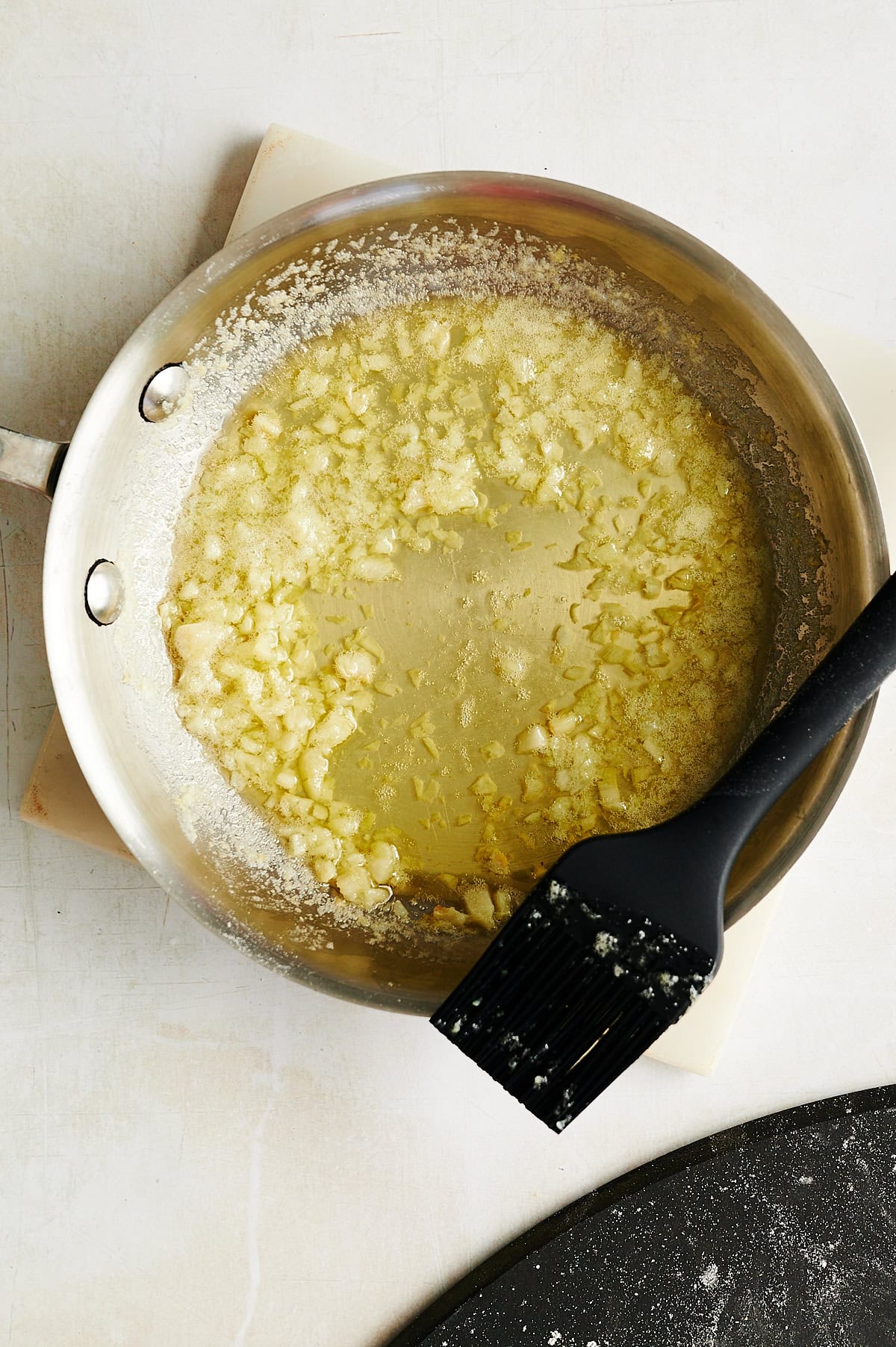 A stainless steel pan containing melted butter and chopped garlic sits on a white surface with a black silicone brush on the edge of the pan.