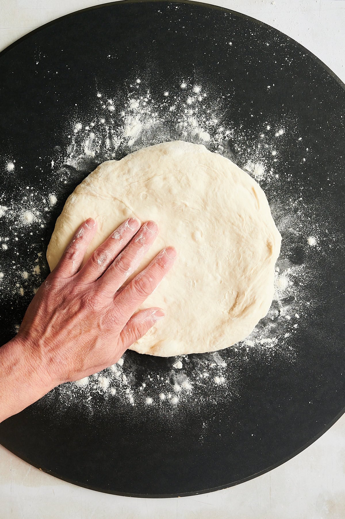 A hand presses down on a round piece of dough, which is sprinkled with flour and resting on a black surface also dusted with flour.