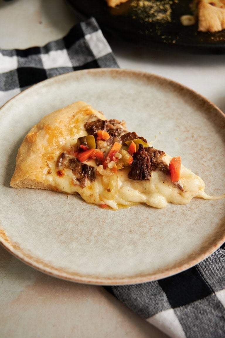 A slice of pizza with melted cheese, shredded meat, and chopped peppers on a beige plate, placed on a black and white checkered cloth napkin.