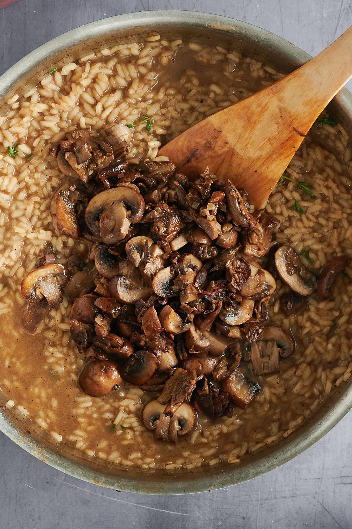 A pot of mushroom risotto is being stirred with a wooden spatula. The dish contains creamy rice and sliced mushrooms, appearing in a thick, brown sauce. The image features the risotto from a top-down perspective against a gray background.