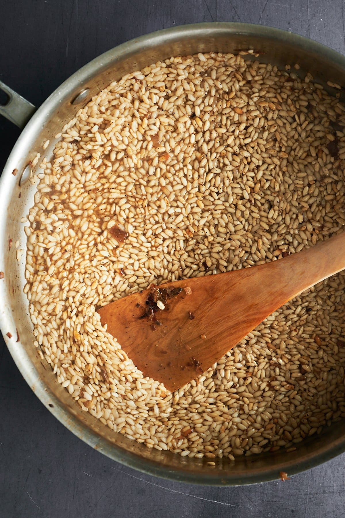 A large metal pan is filled with uncooked rice or a similar grain. A wooden spatula rests inside, angled diagonally from the bottom left to the center. The grains are light brown, and the pan sits on a dark surface.