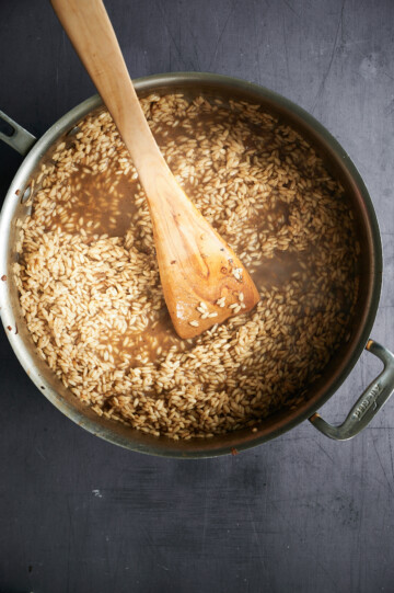 A pot of risotto is shown, with rice grains cooking in broth. A wooden spoon rests in the pot, stirring the rice. The pot is placed on a dark surface.