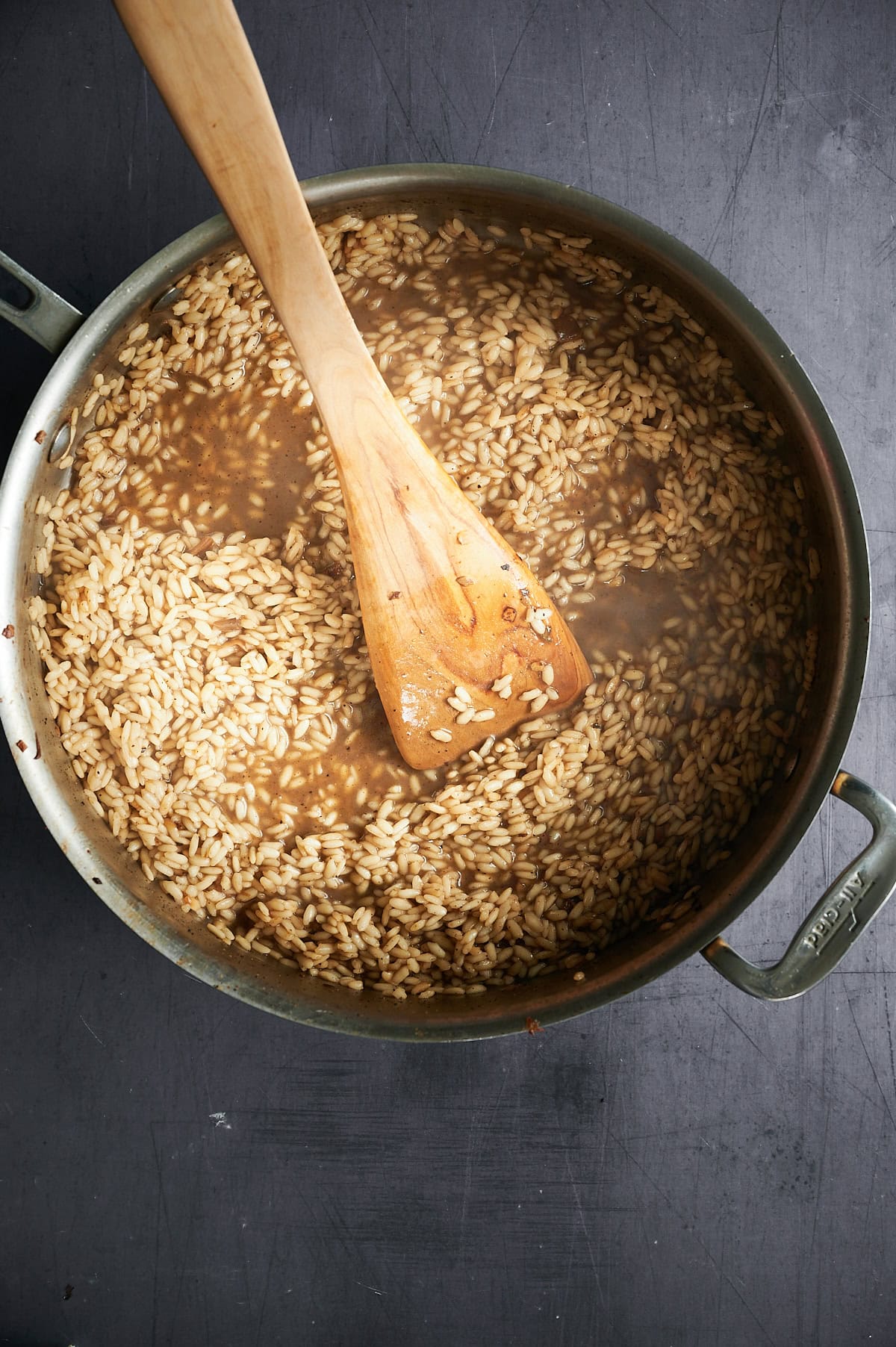 A pot of risotto is shown, with rice grains cooking in broth. A wooden spoon rests in the pot, stirring the rice. The pot is placed on a dark surface.