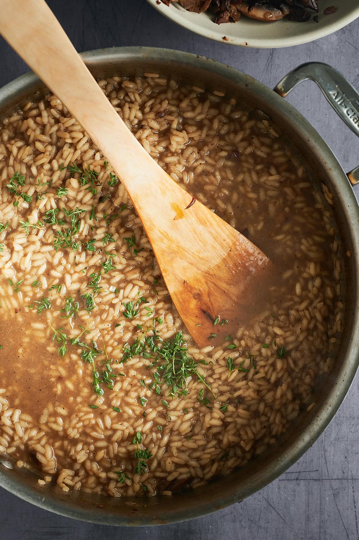 A pot of risotto with a wooden spoon resting inside. The rice is partially submerged in a brown broth and garnished with fresh green herbs. The pot is on a gray surface, and a small portion of mushrooms is visible at the top edge.
