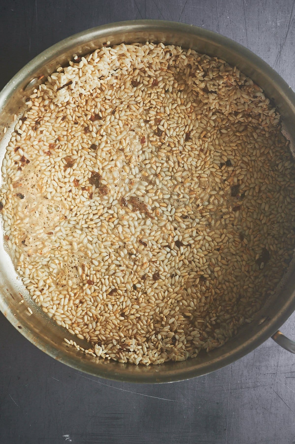Close-up of a pot filled with uncooked rice grains spread evenly across the bottom. The grains are pale brown and scattered with bits of seasoning. The pots metallic edge is visible, resting on a dark gray surface.