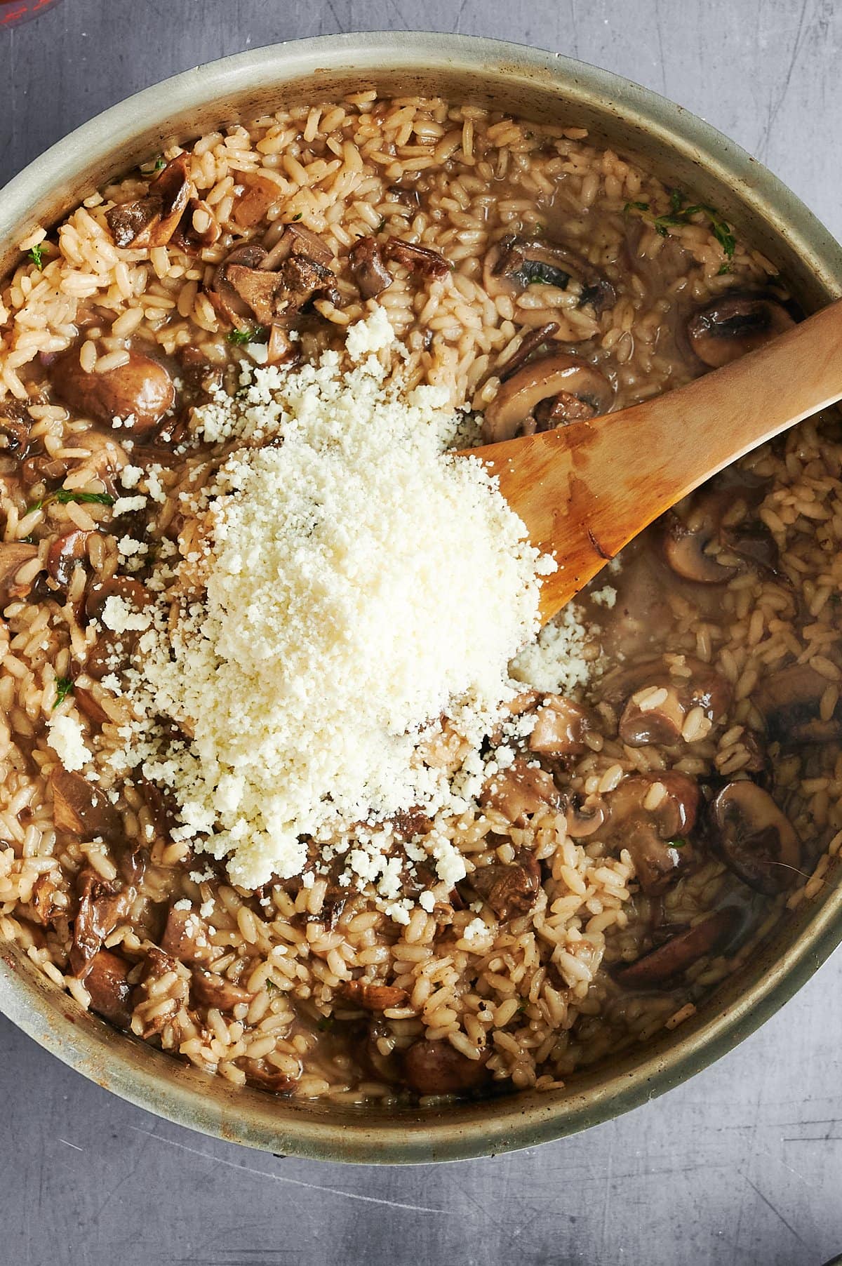 A large skillet contains risotto with visible mushrooms and rice. A wooden spoon is in the pan, and there is a pile of grated cheese on top, ready to be mixed in. The background surface is gray.