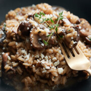 A plate of mushroom risotto topped with grated cheese and garnished with a sprig of thyme. A fork is resting on the risotto, set against a dark background.