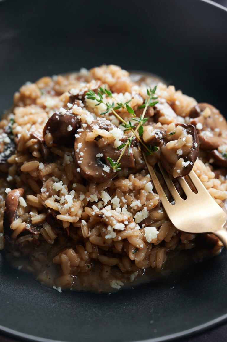A plate of mushroom risotto topped with grated cheese and garnished with a sprig of thyme. A fork is resting on the risotto, set against a dark background.