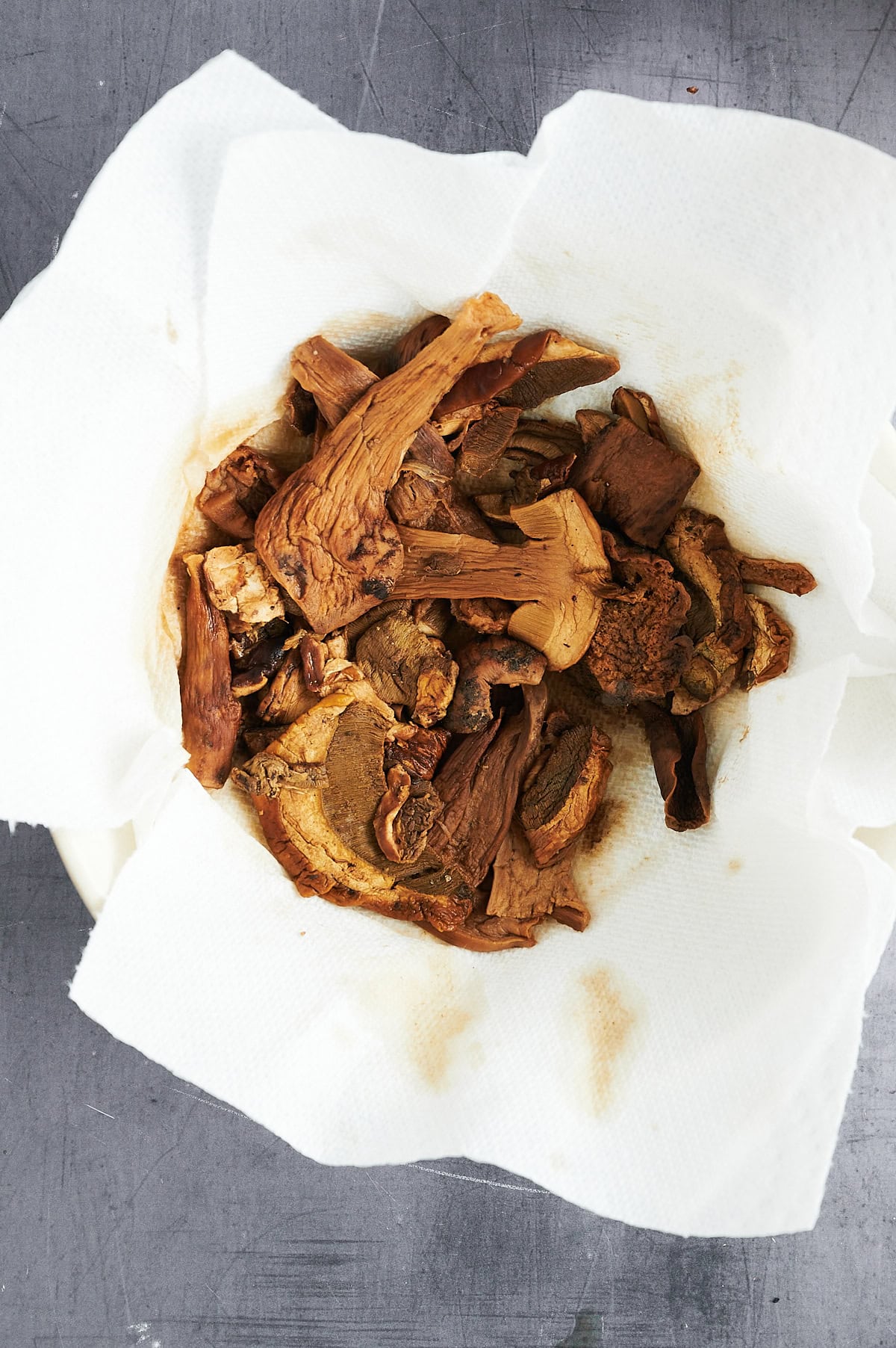 A bowl of dried mushrooms sits on a paper towel, placed on a gray surface. The mushrooms are brown and irregularly shaped, showing signs of dehydration.