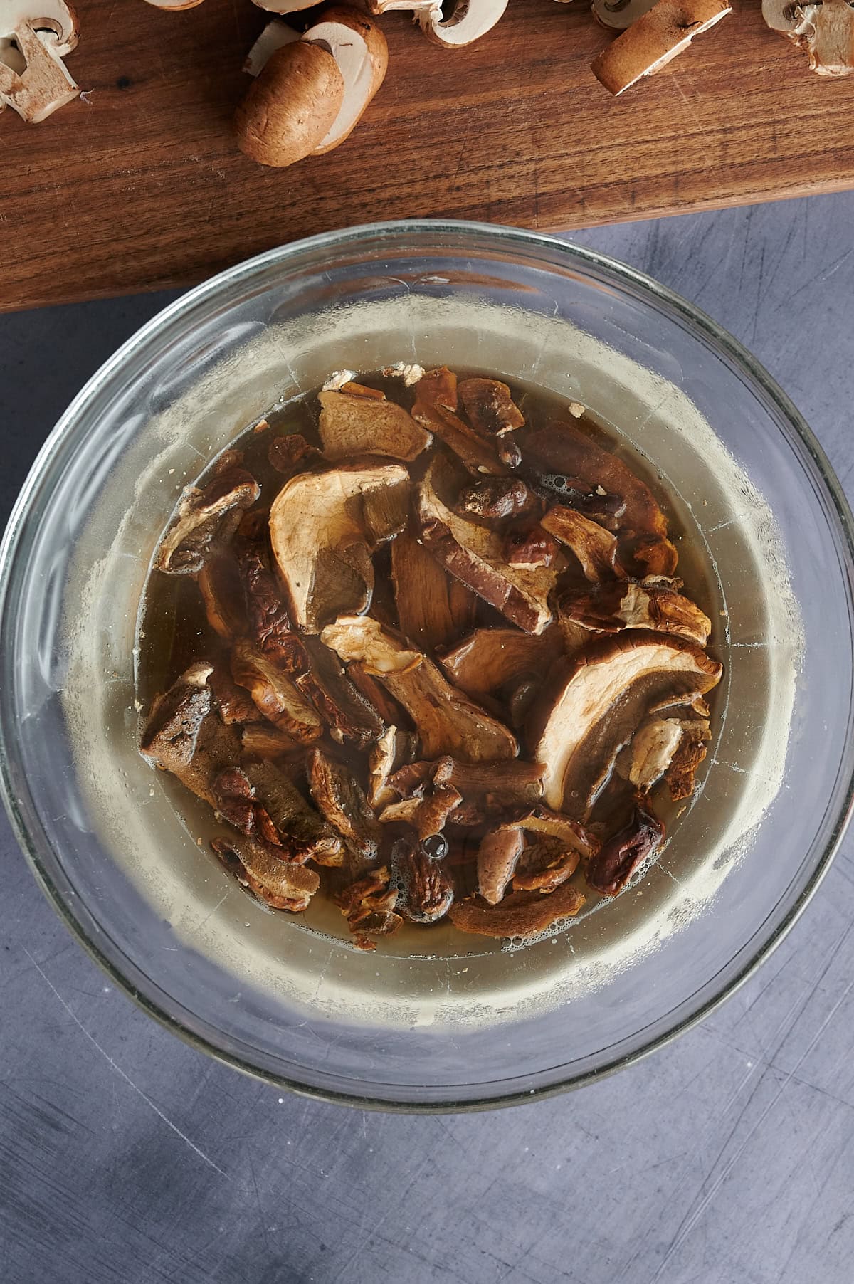 A glass bowl filled with sliced mushrooms soaking in water. Above, a wooden cutting board with additional mushroom pieces is visible. The bowl sits on a gray surface.