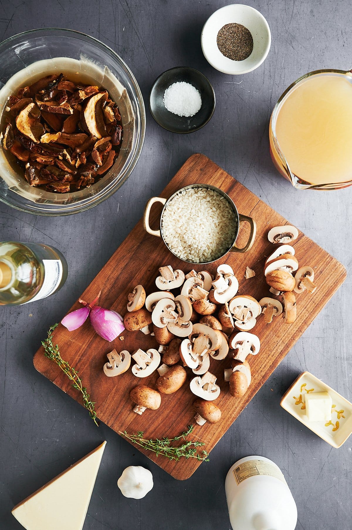 Ingredients are arranged on a gray table: sliced mushrooms, rice in a small pot, shallots, thyme, and butter on a wooden cutting board, along with bowls of soaked mushrooms, salt, and pepper. A cup of broth and a small bottle of white wine are nearby.