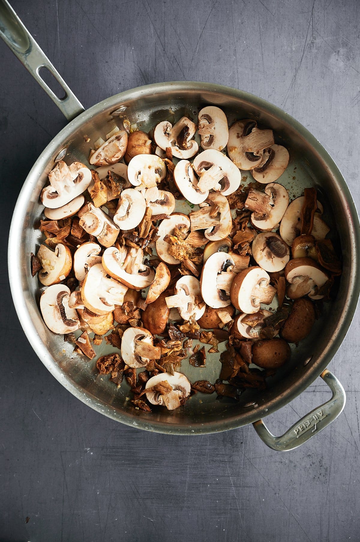 A metal pan on a dark surface contains a mixture of sliced and whole mushrooms. The mushrooms vary in size and are primarily in shades of brown and white. Some appear cooked while others look raw. The pan has two handles.