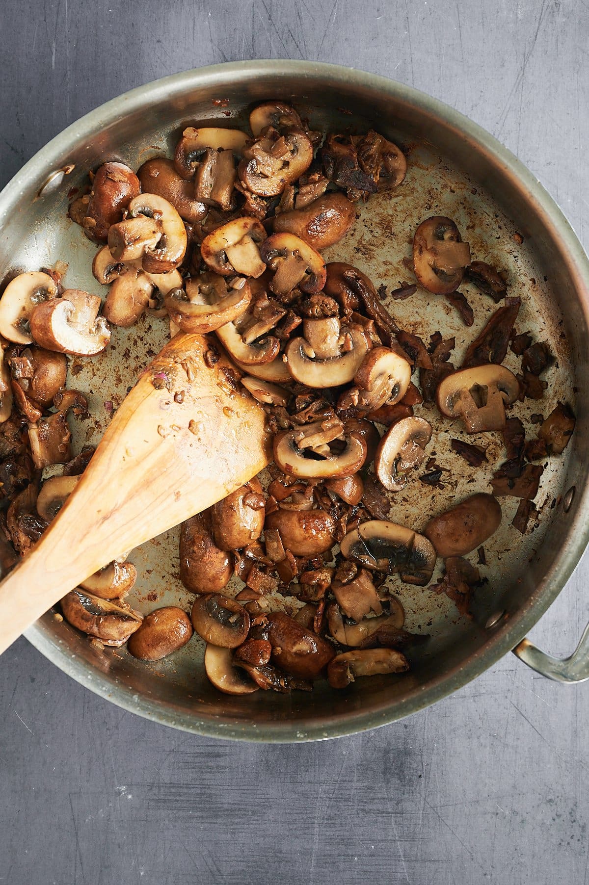A metal frying pan contains sliced and whole cooked mushrooms. A wooden spatula rests inside the pan, partially covered by the mushrooms. The background is a textured gray surface.