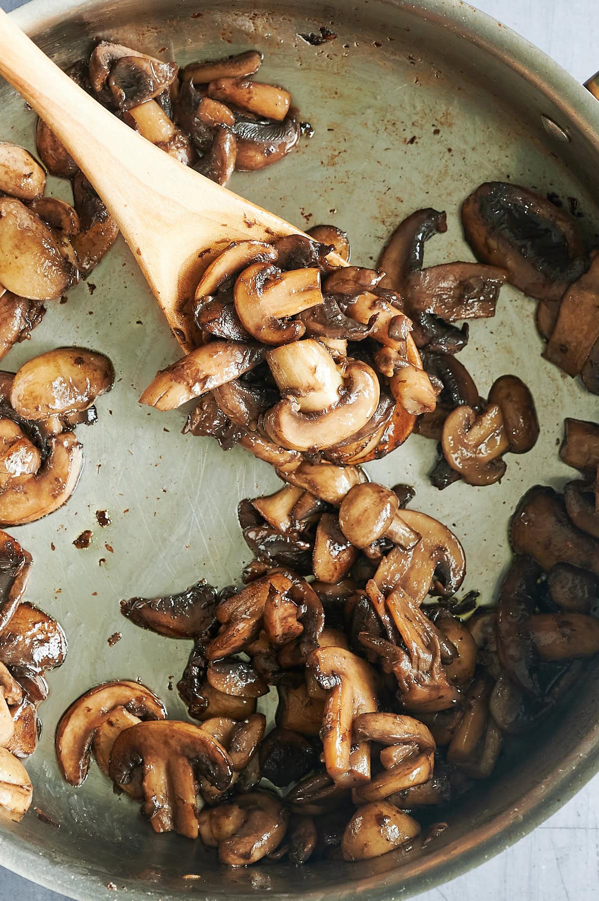 Sliced mushrooms being sautéed in a pan, stirred with a wooden spoon. The mushrooms are browned and appear cooked, with visible pieces spread across the surface of the pan.