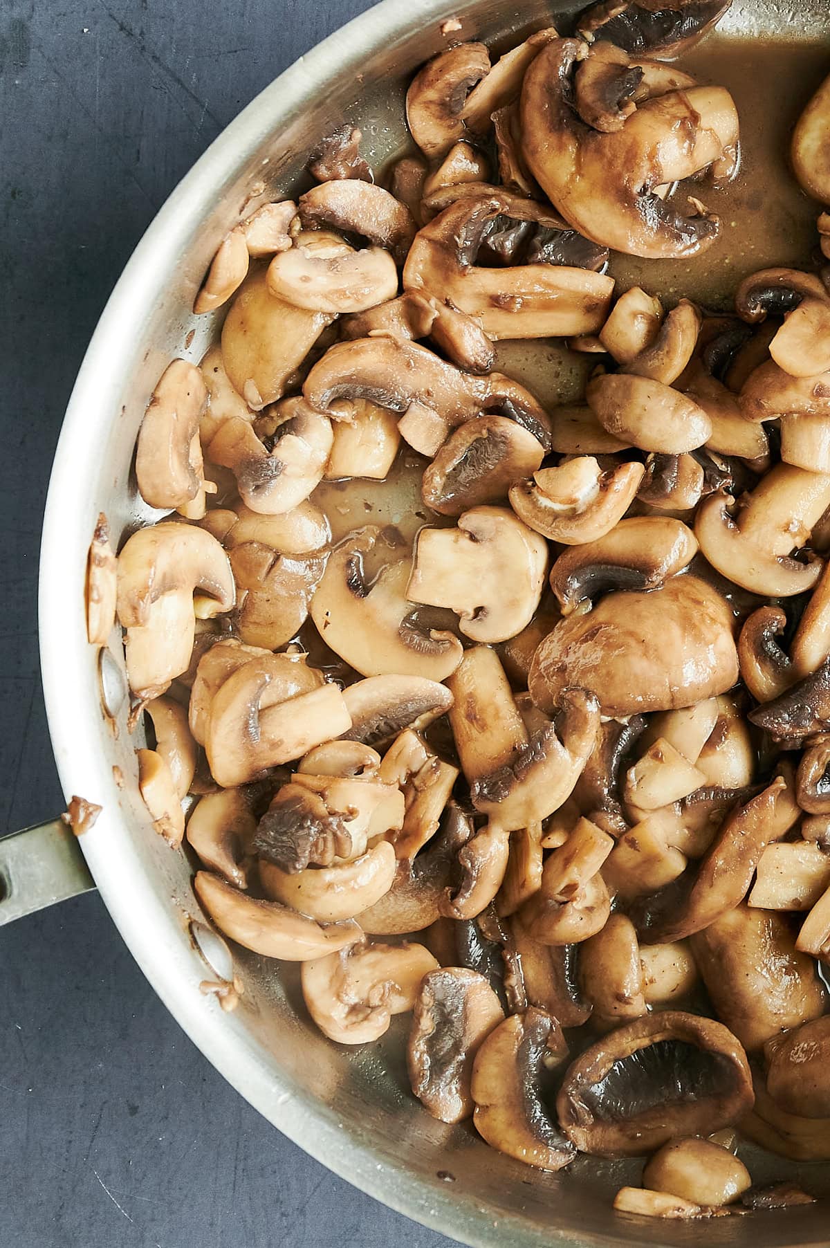 A stainless steel pan filled with sliced mushrooms being sautéed, showing a mix of browned and lightly cooked pieces on a dark countertop.