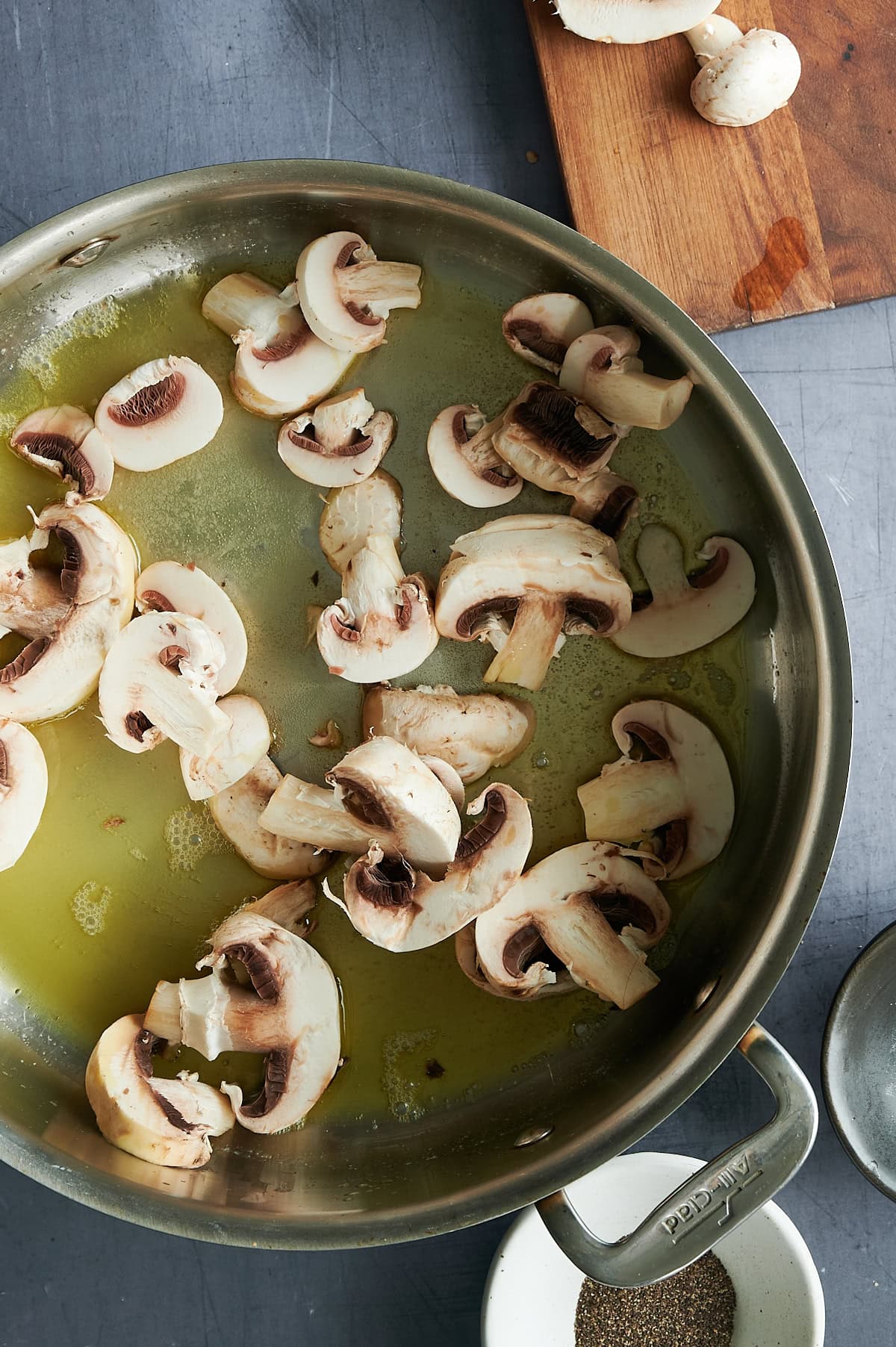 A stainless steel pan with sliced mushrooms cooking in oil is on a gray surface.
