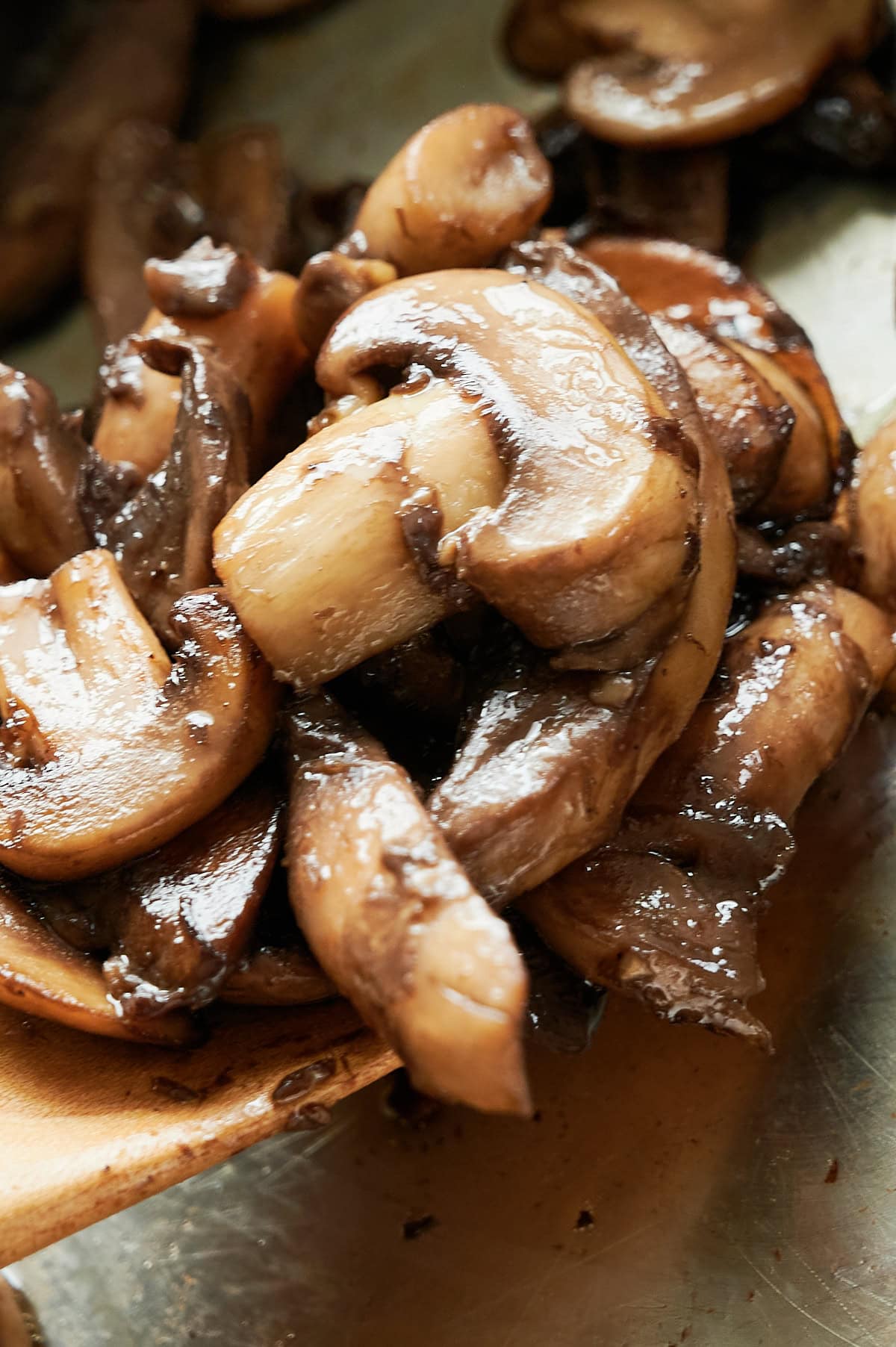 Close-up of sliced, sautéed mushrooms being scooped with a wooden utensil. The mushrooms are golden brown and appear glossy from cooking.