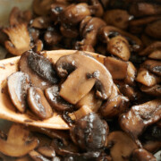Close up of sliced mushrooms being sautéed in a pan, with a wooden spoon lifting some of the cooked mushrooms. The mushrooms appear browned and glossy from cooking.