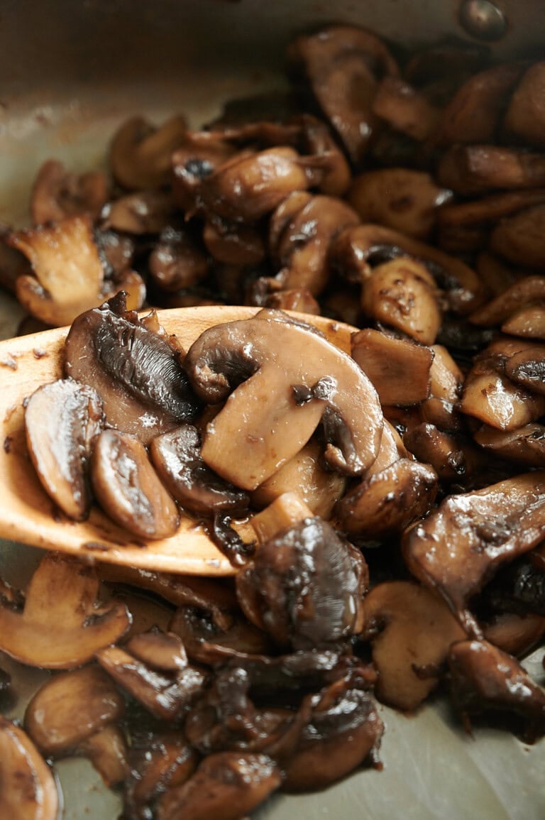 Close up of sliced mushrooms being sautéed in a pan, with a wooden spoon lifting some of the cooked mushrooms. The mushrooms appear browned and glossy from cooking.