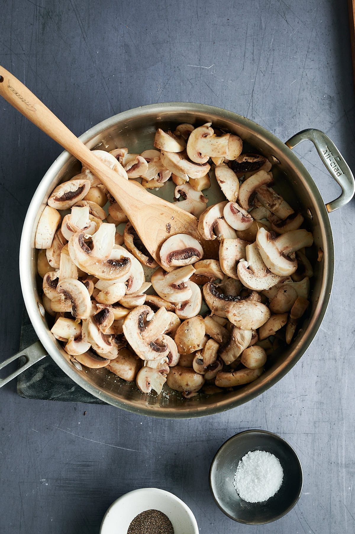 A stainless steel pan filled with sliced mushrooms being stirred with a wooden spoon on a gray surface, with small bowls of salt and pepper nearby.
