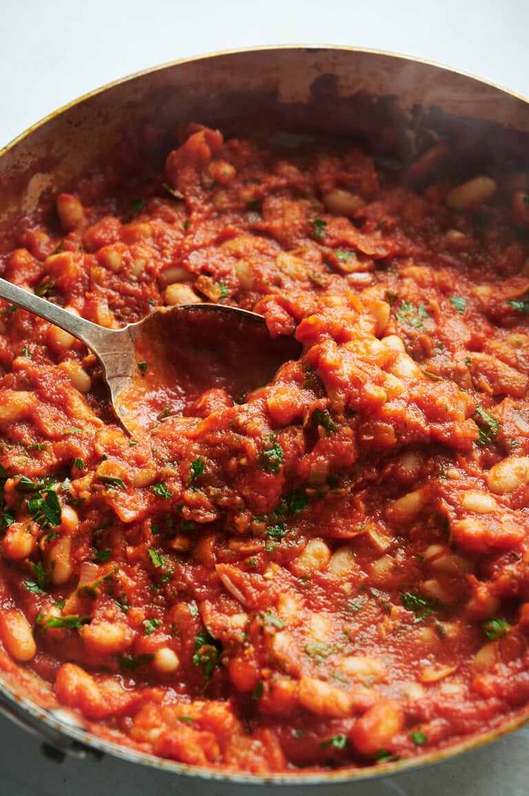 A close-up of a pan filled with a tomato-based pan of cannellini beans and chopped herbs, with an antique spoon.
