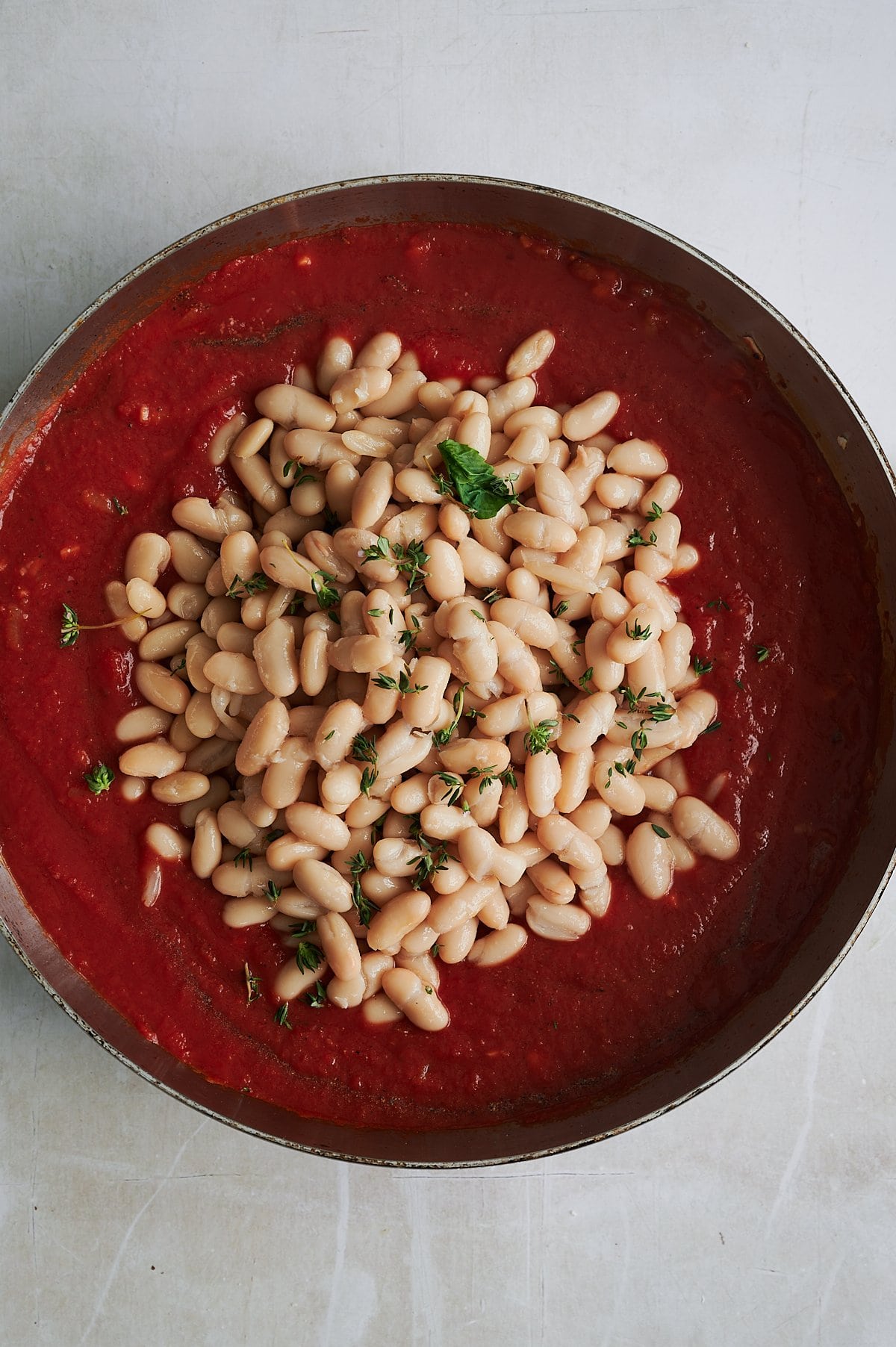 A pan filled with tomato sauce, topped with cooked cannellini beans and garnished with fresh herbs, sits on a light-colored surface.