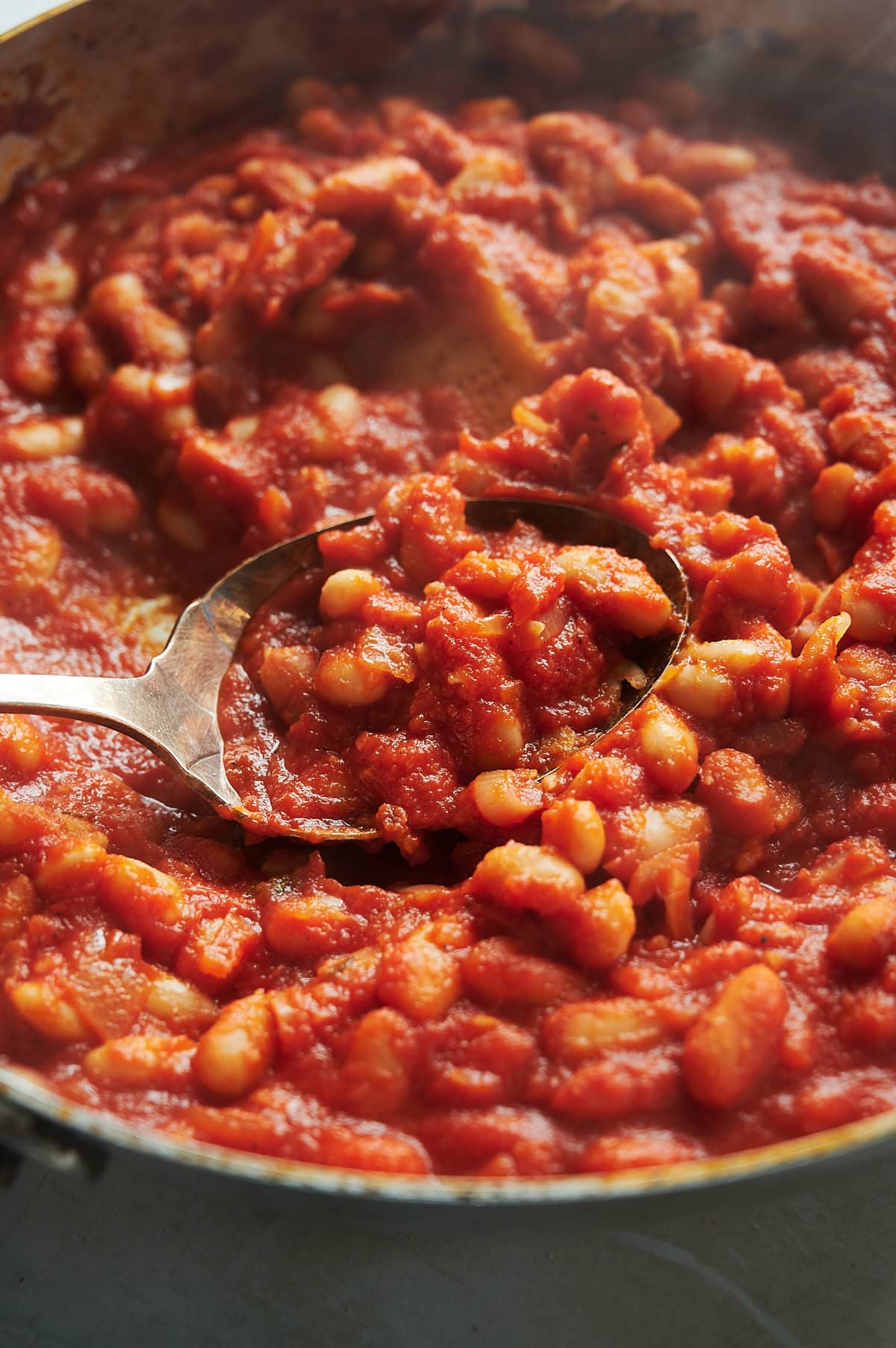 A close-up view of a pan filled with cannellini beans in a thick tomato sauce, with a metal serving spoon scooping some of the beans.