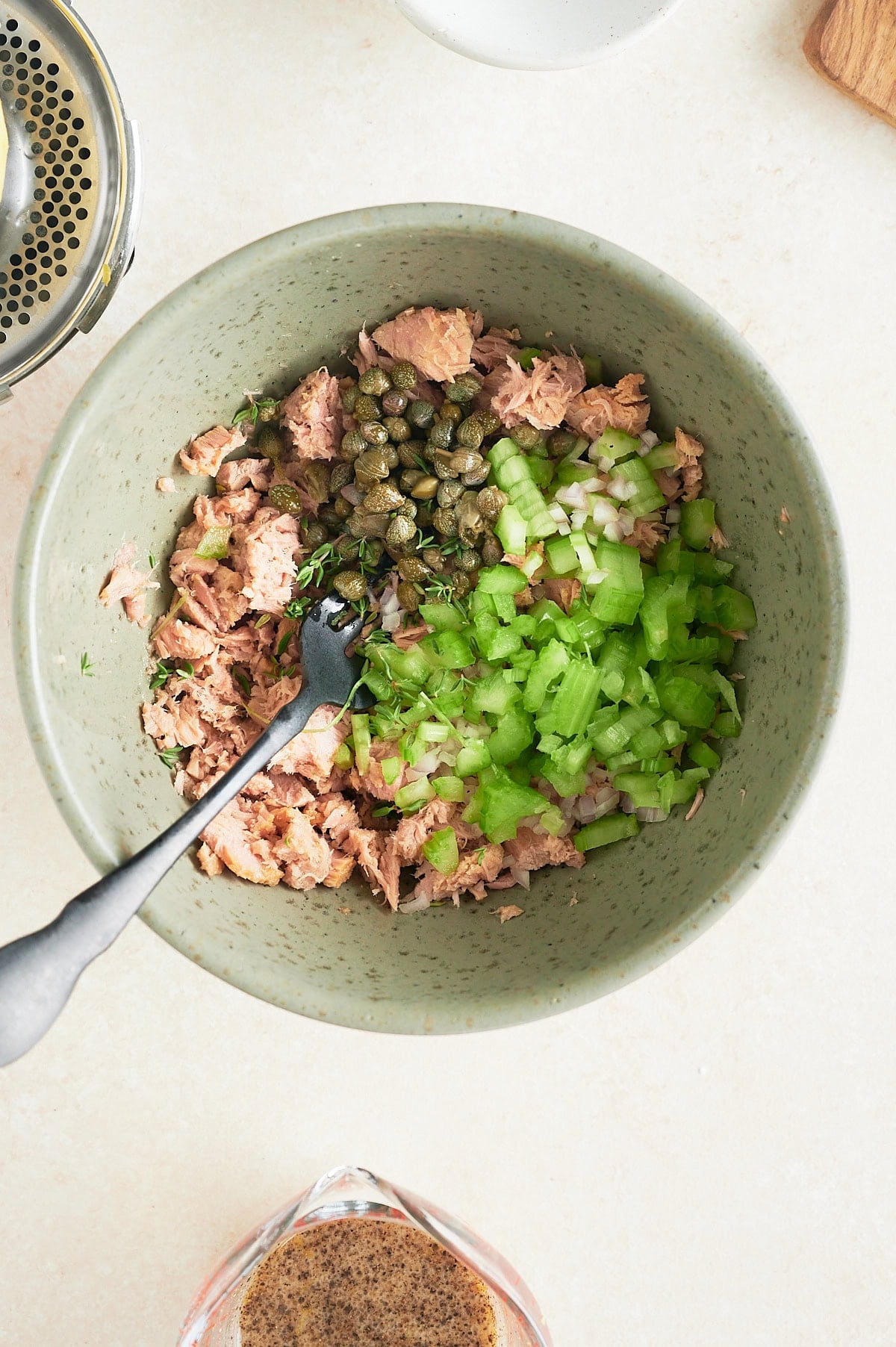 A bowl containing flaked tuna, chopped green celery, chopped white onion, and capers with a fork resting inside; a measuring cup with dressing and a juicer are nearby on a light surface.