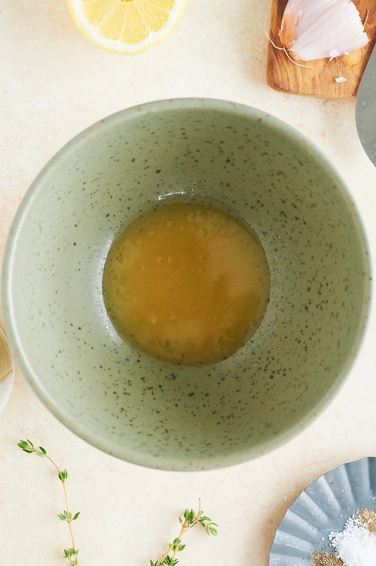 A green speckled bowl contains a small amount of yellow liquid—perfect for preparing Italian tuna salad. Surrounding it are a lemon wedge, clove of garlic on a wooden board, thyme sprig, and a plate with salt and pepper.